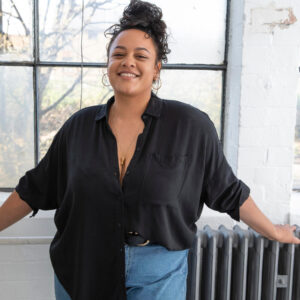 Selbstbewusste Frau in einer schwarzen Bluse steht fröhlich vor einem Fenster