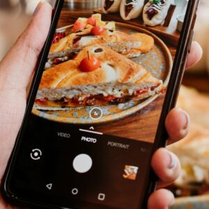 Zu sehen ist eine hand, die ein Smartphone hält, auf dessen Display gerade ein Gericht fotographiert wird-