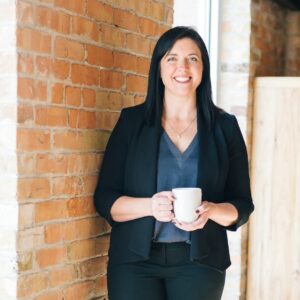 Frau im schwarzen Anzug mit dunklen Haaren hält Tasse in der hand und lacht