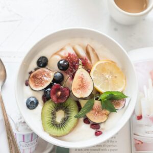 Eine Joghurt Bowl mit Früchten steht zusammen mit einem Buch und einer Tasse Kaffee auf einer weißen Platte