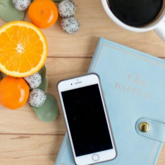 Auf einer Tischplatte liegt ein blaues Notizbuch, darauf steht eine Tasse Kaffe und ein Handy. Daneben liegt Obst.