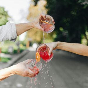 Vier Hände halten verschiedene Gläser mit roter Limonde und schütten diese aus, sodass die Limonde über die anderen Gläser darunter fließt.