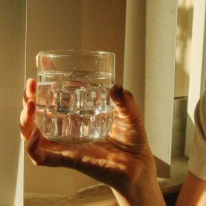 Hand hält ein Glas Wasser vor einem Fenster in warmen Licht.