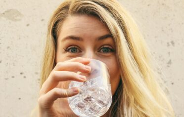 Frau mit blonden Haaren trinkt ein Glas Wasser und guckt in die Kamera.