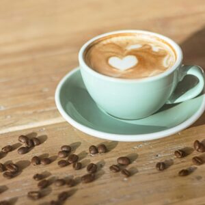 Kaffee in mint farbener Tasse auf einer Holzplatte, auf der Kaffeebohnen verteilt sind.