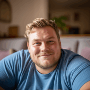 Mann mit blonden Haaren im blauen T-Shirt sitzt lächenld auf einem Sofa.