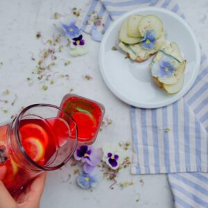 Auf einer weißen Tischplatte steht ein Teller mit Obst auf einem gestreiften Handtuch. Daneben schenkt eine Hand rote Limonade mit Früchten auf einer Karaffe in ein Glas.