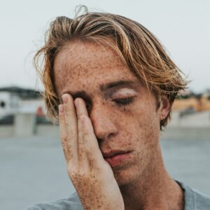 Ein gestresst wirkender junger Mann mit Sommersprossen fässt sich ins Gesicht.