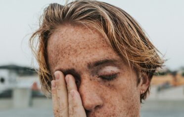 Ein gestresst wirkender junger Mann mit Sommersprossen fässt sich ins Gesicht.