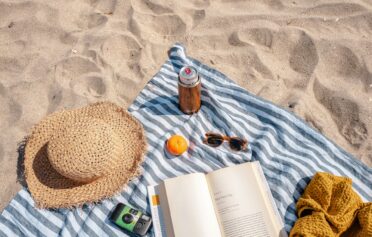Auf einem gestreiften Handtuch am Strand liegen ein Sonnenhut, eine Sonnenbrille und ein Buch.