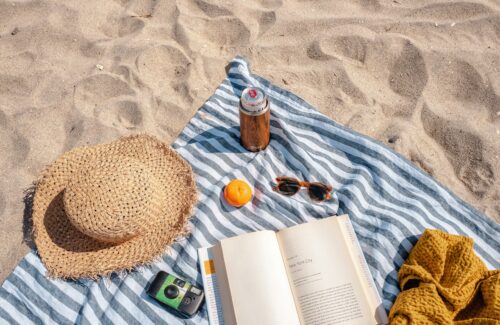 Auf einem gestreiften Handtuch am Strand liegen ein Sonnenhut, eine Sonnenbrille und ein Buch.