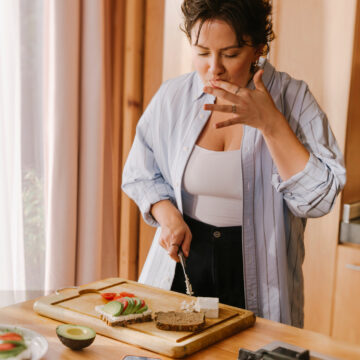 Person bereitet in einer warm beleuchteten Küche ein Brot mit Avocado, Tomaten und Frischkäse zu und probiert dabei etwas von den Fingern.