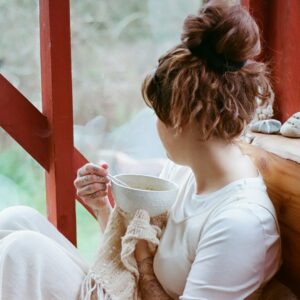 Eine junge Frau sitzt mit einer Tasse Tee im Wintergarten und schaut nach draußen.