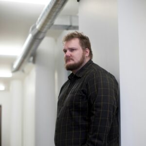 Ein plus size Mann lehnt an einer Wand und guckt in den Raum.