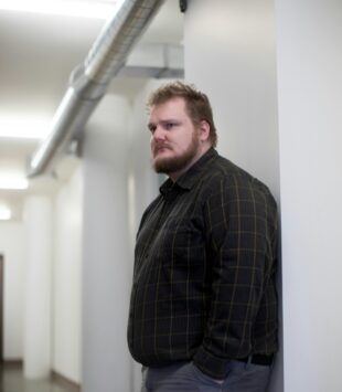 Ein plus size Mann lehnt an einer Wand und guckt in den Raum.