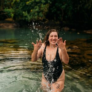 Eine junge plus size Frau steht im Badeanzug in einem See und spritzt Wasser hoch.