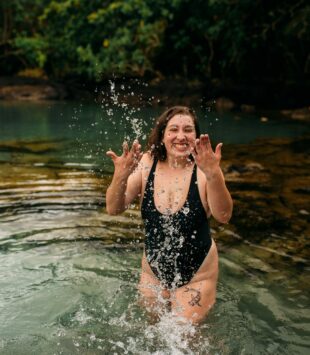 Eine junge plus size Frau steht im Badeanzug in einem See und spritzt Wasser hoch.