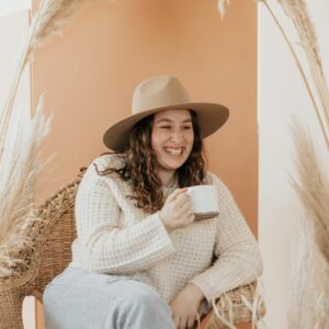 Eine junge plus size Frau mit Hut sitzt mit einer Tasse Kaffee lachend in einem Sessel.