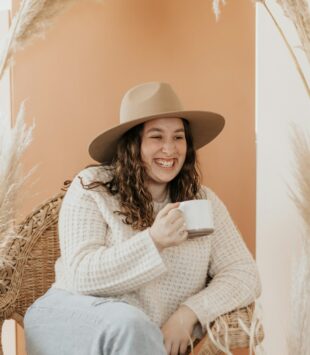 Eine junge plus size Frau mit Hut sitzt mit einer Tasse Kaffee lachend in einem Sessel.