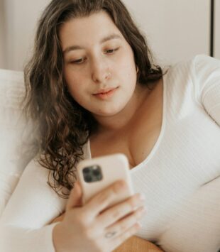 Eine junge plus size Frau liegt auf ihrem Bett und schaut auf ihr Handy.