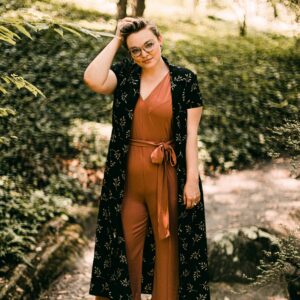 Eine junge Frau steht selbstbewusst in einem grünen Garten und schaut in die Kamera.