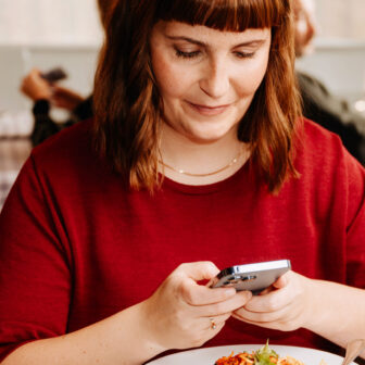 Eine Frau sitzt in einem Restaurant, trägt ein weinrotes Oberteil und schaut lächelnd auf ihr Smartphone. Vor ihr steht ein Teller mit Spaghetti und Basilikum.