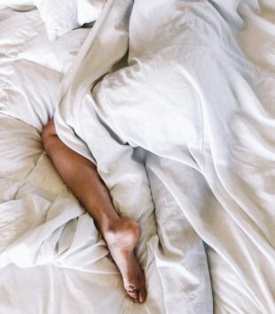 Eine Person liegt schlafend im Bett und ist bedeckt mit der Bettdecke, nur ein Bein guckt heraus.