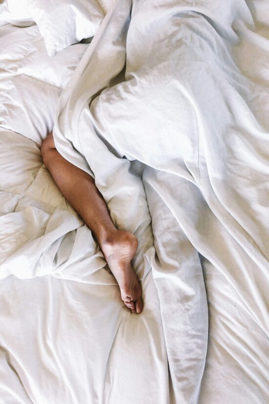Eine Person liegt schlafend im Bett und ist bedeckt mit der Bettdecke, nur ein Bein guckt heraus.