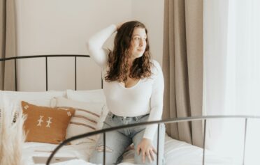 Eine junge Frau kniet auf einem Bett und blickt aus dem Fenster.