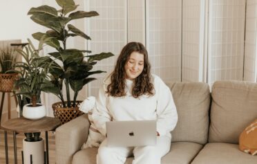 Eine junge plus size Frau sitzt auf ihrem Sofa mit ihrem Laptop auf dem Schoß und schaut lächelnd auf den Bildschirm.