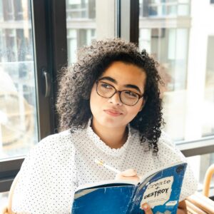 Eine junge Frau sitzt mit einem Buch vor einem Fenster und blickt nachdenklich zur Seite.