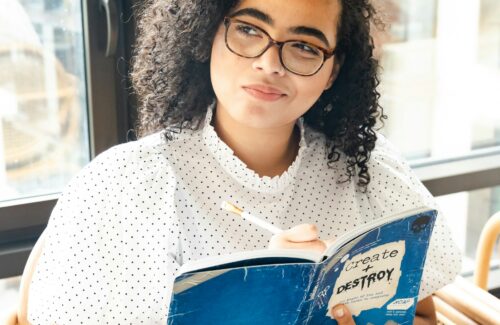 Eine junge Frau sitzt mit einem Buch vor einem Fenster und blickt nachdenklich zur Seite.
