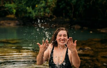 Eine junge plus size Frau in einem schwarzen Badeanzug steht in einem See und spritzt fröhlich lächelnd Wasser in die Luft.