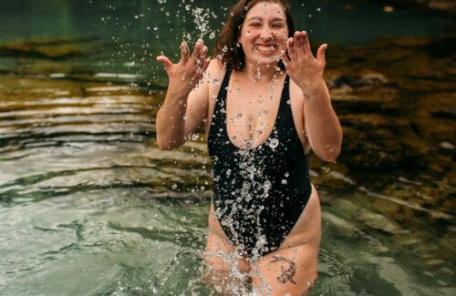 Eine junge plus size Frau in einem schwarzen Badeanzug steht in einem See und spritzt fröhlich lächelnd Wasser in die Luft.