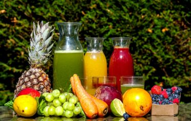 Frisches Obst und Gemüse und drei Karaffen mit frischem Saft und Gläsern sind auf einem Gartentisch drapiert.