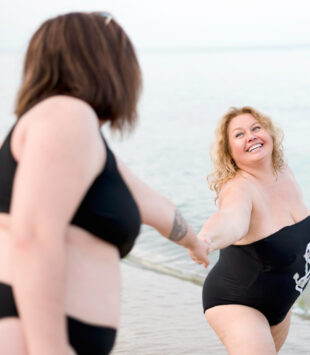 Eine plus size Frau steht im Badeanzug am Strand und hält eine andere Frau an der Hand.