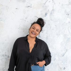 Eine junge curvy Frau steht lässig vor einer Wand und lächelt in die Kamera.