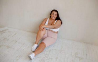 Eine junge plus size Frau sitzt in workout Kleidung auf dem Boden und lächelt.