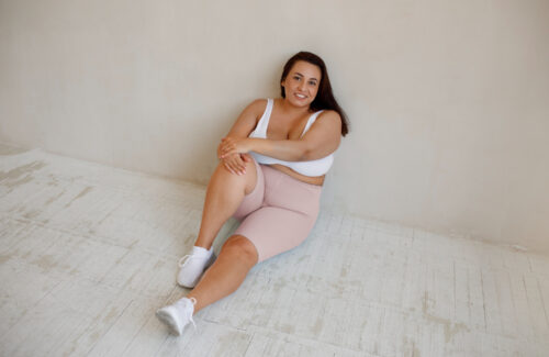 Eine junge plus size Frau sitzt in workout Kleidung auf dem Boden und lächelt.