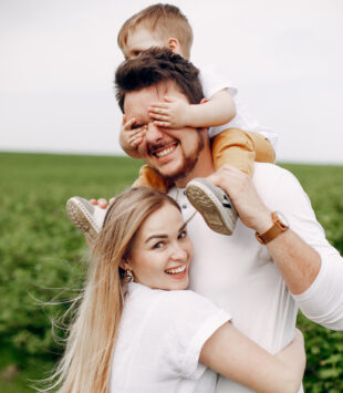 Ein Bild einer jungen Familie, auf dem die Frau ihren Mann umarmt, der auf den Schulter ihr Kind trägt. Alle lachen fröhlich.