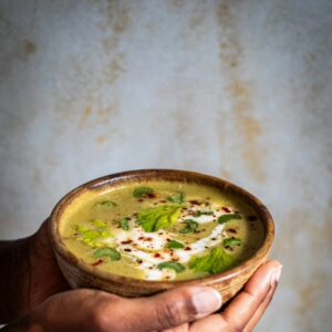 Hände halten eine Bowl mit grüner Gemüsesuppe in der Hand.