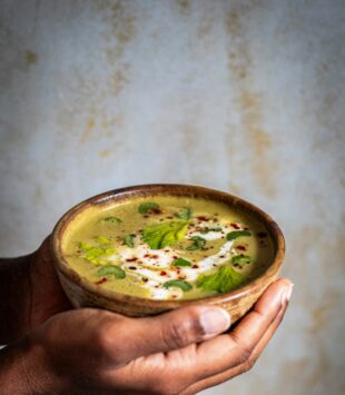 Hände halten eine Bowl mit grüner Gemüsesuppe in der Hand.