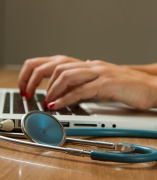 Eine Nahaufnahme von Händen, die auf einem Laptop tippen und daneben liegt ein Stetoskop.