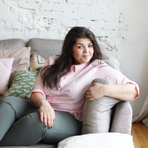 Eine junge Frau sitzt auf einem Sofa und guckt seitlich lächelnd zur Kamera, während sie sich mit ihrem linken Arm auf der Sofalehne abstützt.