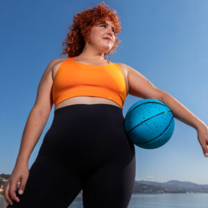 Eine kurvige Frau von unten abgelichtet steht vor einem See, der Himmel ist blau. Sie trägt Sportkleidung und hat unter ihrem linken arm einen Basketball klemmen.