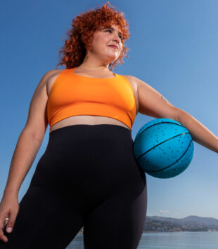Eine kurvige Frau von unten abgelichtet steht vor einem See, der Himmel ist blau. Sie trägt Sportkleidung und hat unter ihrem linken arm einen Basketball klemmen.