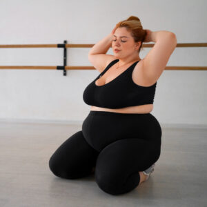 Eine junge Frau in schwarzer Sportbekleidung kniet auf dem Boden eines Sportraumes und hat ihre Arme hinter dem Kopf verschränkt.