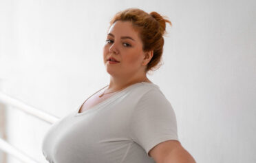 Eine curvy Frau in Sportkleidung steht angelehnt an einer Barrestange.