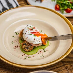 Ein Teller mit einer kleinen Schnitte Brot auf einem Kräuteröl mit einem pochierten Ei darauf, welches gerade mit einem Messer aufgeschnitten wird, sodass das flüssige Eigelb herausläuft.