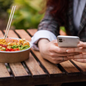 Eine junge Frau sitzt draußen an einem Tisch mit einer Salatbwol und tippt nebenbei auf ihrem Smartphone.